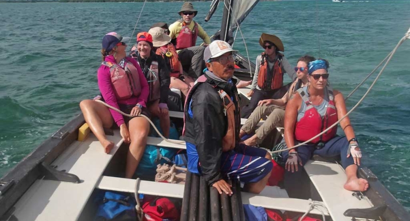 A group of adults wearing lifejackets sit on a sailboat floating on blue water